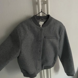 Grå bomberjacka från H&M - Säljer en stilren grå bomberjacka från H&M med ribbade muddar och krage. Jackan har knappar framtill och en diskret bröstficka. Perfekt för dig som gillar enkel och clean look. Kommer ej till användning. Passar XS och S