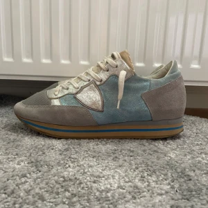Phillipe model skor - Säljer ett par snygga sneakers från Philippe Model i grått och ljusblått med inslag av vitt. Skorna har ovandel i mocka och mesh, vita snören och en platt sula med blå och beige ränder. Perfekta för dig som gillar stilrena och sportiga skor. Skorna är i storlek 40 och passar perfekt till sommaren med ett nypris på cirka 3000 kr!