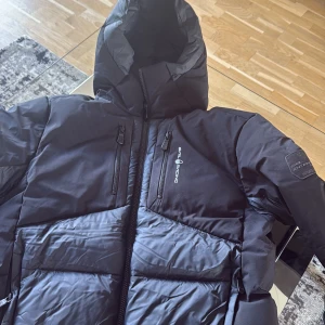 Svart Sail Racing dunjacka med huva - Säljer en svart dunjacka från Sail Racing, modell Antarctica Expedition. Jackan har huva, två bröstfickor med dragkedja och Sail Racing-logga på bröstet samt patch på ärmen. Perfekt för kalla dagar och riktigt snygg streetstil.