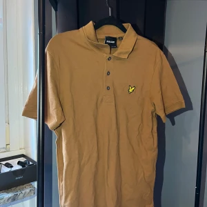 Brun pikétröja från Lyle & Scott - Snygg brun pikétröja från Lyle & Scott med klassisk krage och tre knappar framtill. Tröjan har korta ärmar och den ikoniska gula loggan broderad på bröstet. Tillverkad i mjuk bomull, perfekt för en clean och stilren look.