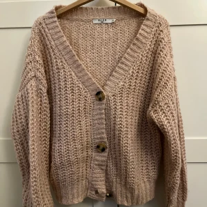 Beige stickad kofta från NA-KD - Supermysig beige stickad kofta från NA-KD med stora knappar framtill och djup v-ringning. Koftan har långärmade ärmar och en loose fit, perfekt att slänga på sig när det är kyligt ute. Passar även xs-s