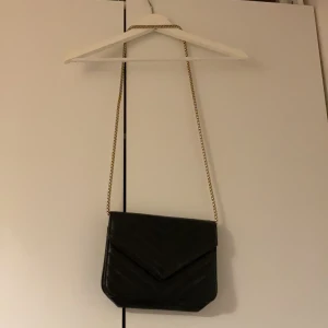 Svart quiltad axelväska med kedja - Snygg svart axelväska med quiltad design och guldfärgad kedjerem. Väskan har ett kuvertformat lock och är tillverkad i skinnimitation. Perfekt för dig som vill ha en stilren och trendig accessoar.