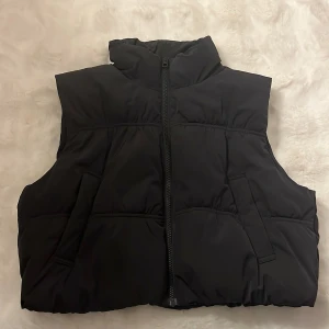 Svart dunväst från H&M Divided - Svart dunväst från H&M Divided i storlek S. Västen har hög krage, två fickor och stängs med dragkedja framtill. Perfekt för lager-på-lager och har en enkel, stilren look som funkar till många outfits.