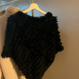 Svart pälsponcho från Soyaconcept - Säljer en svart poncho från Soyaconcept i äkta kaninpäls och akryl. Den har en fluffig och lyxig känsla, perfekt för att ge din outfit en extra edge. Ponchon har en asymmetrisk form och är supermjuk och varm.