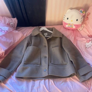 Ljusbrun kappa från Kappahl - Stilren ljusbrun kappa från Kappahl i ullmix med stora fickor framtill och klassisk krage. Kappan har knäppning med knappar och uppvikta ärmslut för en trendig look. Perfekt för lager-på-lager under höst och vår.