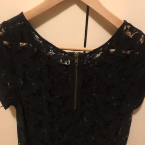 Svart spetslinne med dragkedja - Svart kortärmad klänning i spets med blommigt mönster och rund halsringning. Klänningen har en synlig dragkedja bak och är figurnära i modellen. Perfekt för dig som gillar en stilren och elegant look.