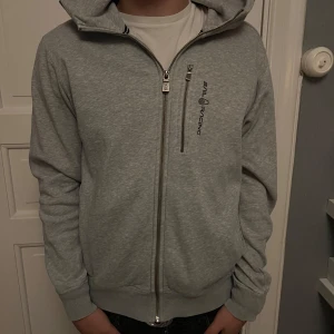 Grå hoodie från Sail Racing - Säljer en stilren grå hoodie från Sail Racing med dragkedja framtill och en extra bröstficka med zip. Tröjan har huva, långa ärmar och ribbade muddar. Perfekt för dig som gillar en clean och sportig look.