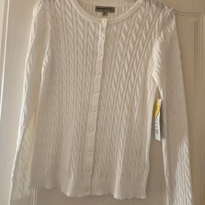 Vit kabelstickad kofta Basic You S - Kabelstickad kofta i offwhite från Basic You, tillverkad i 100% ekologisk bomull. Klassisk rund hals, långa ärmar och knäppning framtill med vita knappar. Perfekt för lager-på-lager och enkel att matcha med allt.