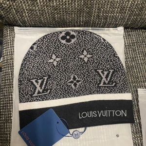 Svart Louis Vuitton mössa med logga - Snygg svart och vit mössa från Louis Vuitton med klassiskt LV-monogram och blommönster. Mössan har en bred vit rand vid kanten och tydlig Louis Vuitton-text framtill. Stickad i mjukt material, perfekt för att lyfta din stil under kyliga dagar.