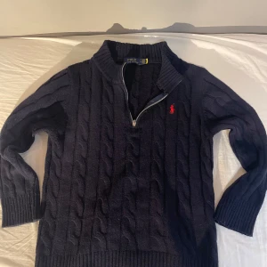 Mörkblå stickad tröja Polo Ralph Lauren - Kabelstickad mörkblå tröja från Polo Ralph Lauren med halv dragkedja och röd broderad logga på bröstet. Tröjan har hög krage och långa ärmar, perfekt för kyliga dagar. Klassisk och stilren design som funkar till det mesta.