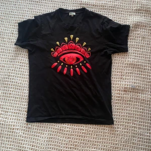 Svart Kenzo t-shirt med ögontryck - Svart t-shirt från Kenzo med ikoniskt ögontryck i rött och gult på bröstet. Klassisk passform och rund hals. Tillverkad i mjuk bomull, perfekt för dig som vill sticka ut med en cool och unik look.