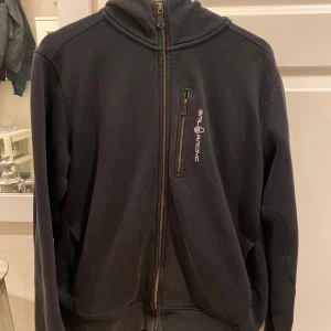 Svart hoodie från Sail Racing - Svart hoodie från Sail Racing med hel dragkedja framtill och en extra bröstficka med dragkedja. Använd men bra skick! Köparen står för frakt!