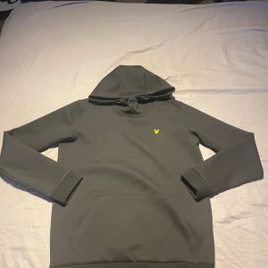 Svart hoodie från Lyle & Scott - Svart hoodie från Lyle & Scott med klassisk gul logga på bröstet. Tröjan har huva med snörning och en stor magficka framtill. Tillverkad i mjukt material, perfekt för chill dagar. Långärmad och stilren design som funkar året runt.