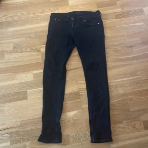 Svarta jeans från Nudie Jeans, W31 L32 - Säljer ett par svarta jeans från Nudie Jeans i storlek W31 L32. Jeansen har klassisk femficksdesign, svarta sömmar och diskret logga på bakfickan. Modellen är slim och raka ben, tillverkade i mjukt bomullstyg med stretch för extra komfort.
