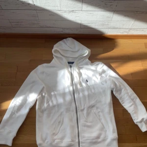 Vit hoodie från Polo Ralph Lauren - Vit hoodie från Polo Ralph Lauren med dragkedja och klassisk broderad logga på bröstet. Tröjan har huva med snörning, två fickor framtill och är tillverkad i mjuk bomull. Perfekt för en chill och stilren look.