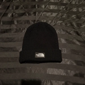 Svart ribbad mössa The North Face - Svart ribbstickad mössa från The North Face med uppvikt kant och klassisk logotyp framtill. Perfekt för kalla dagar och enkel att matcha med olika outfits.