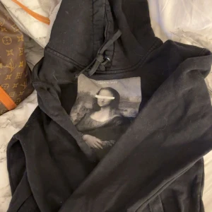 Svart hoodie med Mona Lisa-tryck från the cool elephant - Svart hoodie med snörning i huvan och ett stort tryck av Mona Lisa på bröstet. Tröjan har långa ärmar och en klassisk passform, perfekt för dig som gillar hoodies och konstinspirerade plagg. Pris går alltid att diskutera!!🤍