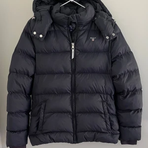 Svart pufferjacka från Gant med huva - Svart vinterjacka från Gant i nyskick, den har bara använts nån enstaka gång. Den är i storlek 170 och passar även XS/S. Det är en unisex så den passar både tjejer och killar.  Modellen är 170cm lång och har S i storlek.