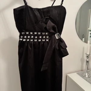 Svart klänning med rosett och stenar - Snygg svart klänning med axelband, plisserad överdel och stor rosett i midjan. Midjan är dekorerad med glittriga stenar och en stor dekorativ sten. Perfekt för dig som vill sticka ut med en elegant look.