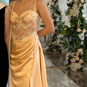 Champagnefärgad långklänning med spets - Superfin långklänning i champagnefärg med glittriga paljettdetaljer och broderad spets upptill. Klänningen har smala axelband, markerad midja och draperad kjol i glansigt satinliknande material. Snygg slits på sidan och transparenta partier vid midjan för extra wow-känsla.