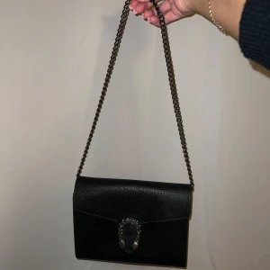 Svart axelväska med kedja och spänne - Stilren svart axelväska i skinn med kedjerem och snyggt silverfärgat spänne framtill. 