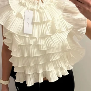 Vit volangblus med plisserade lager - Supersöt vit blus med flera plisserade volanglager och breda ärmar. Blusen har en luftig och romantisk känsla med hög halsringning och är perfekt för dig som vill sticka ut med en unik topp.