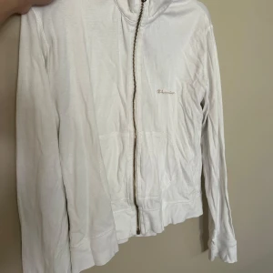 Vit hoodie från Champion med dragkedja - Vit hoodie från Champion med hel dragkedja i metall, broderad logga på bröstet och känguruficka framtill. Klassisk modell med huva och långa ärmar, tillverkad i mjuk bomull. Perfekt för chill dagar eller när du vill ha en clean look.