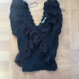 Svart spetsig blus med volanger🫶🏼 - Supersöt svart blus med djup v-ringning och stora volanger framtill. Toppen är stickad med spetsdetaljer och har en croppad passform. Perfekt för dig som gillar romantisk och trendig stil. Säljs utan ärmar och med bred resår nertill🌟.