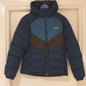 Blå pufferjacka från Urberg, S - Snygg pufferjacka från Urberg i olika blå nyanser med mörkgrå detalj över bröstet. Jackan har huva, dragkedja framtill och två fickor. Perfekt för kalla dagar och har en sportig look med quiltad design.