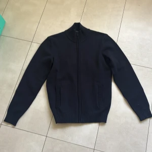 Mörkblå stickad kofta med dragkedja - Snygg mörkblå stickad kofta med hel dragkedja och ribbade detaljer vid krage, fickor och nederkant. Perfekt att slänga på sig när det är lite kyligt. Passar dig som gillar enkel och stilren look.