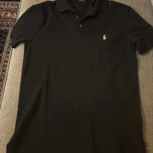 Svart pikétröja Polo Ralph Lauren M - Svart pikétröja från Polo Ralph Lauren i slim fit med klassisk krage och knappar framtill. Ikoniska broderade loggan på bröstet. Tillverkad i stretchig bomullsmix som ger en skön passform. Perfekt för en clean och stilren look. Använt ett par gånger, nypris 1800kr, mitt pris 499!