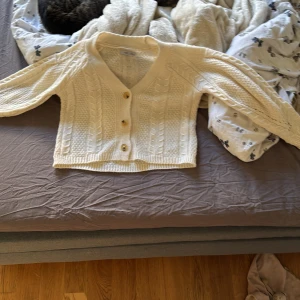 Vit stickad kofta från Hailys - Superfin vit stickad kofta från Hailys med snyggt flätmönster och tre knappar framtill. Koftan har v-ringning, långärmade ärmar och en croppad passform som ger en trendig look. Perfekt att slänga över en topp när det är kyligt ute. Det kommer inte till användning hos mig då den är för liten