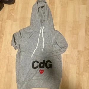 Grå hoodie Comme des Garçons Play S - Grå hoodie från Comme des Garçons Play med vit snörning i huvan och svart CdG-tryck framtill. Ikoniskt rött hjärta med ögon både fram och bak. Mjuk bomullskvalitet och klassisk passform, perfekt för dig som gillar streetstyle.
