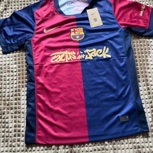 FC Barcelona x Cactus Jack Nike tröja - Exklusiv FC Barcelona x Cactus Jack matchtröja från Nike. Halva tröjan är vinröd och andra halvan marinblå, med gult Nike-märke och klubbmärke på bröstet. Tillverkad i lätt, ventilerande Dri-FIT polyester. Unik Cactus Jack-grafik framtill och UNHCR-logga bak.