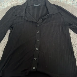 Svart randig tröja  från Lindex - Svart skjorta/tröja  från Lindex i storlek S med diskret randigt mönster. Skjortan har långa ärmar, klassisk krage och knappar hela vägen framtill. Materialet känns mjukt och lätt, perfekt för en chill och stilren look. använt fåtal gånger