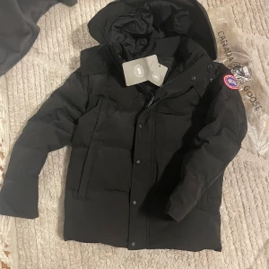 Svart dunjacka från Canada Goose - Säljer en svart dunjacka från Canada Goose med huva och klassisk logga på ärmen. Jackan är vadderad och har dragkedja samt knappar framtill. Medföljer originalpåse och avtagbar pälskant till huvan. Perfekt för kalla vinterdagar.