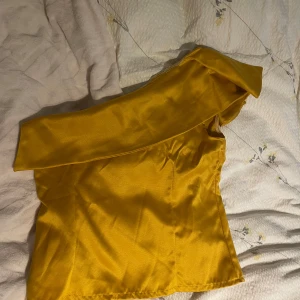 Gul one shoulder topp från DEN-D - Snygg gul topp från DEN-D i glansigt material med en axel och voluminös ärm. Toppen har en cool one shoulder-design och är figursydd för en trendig look. Perfekt för dig som vill sticka ut med färg och form.