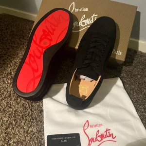 Christian Louboutin svarta sneakers - Säljer ett par svarta sneakers från Christian Louboutin i mocka med klassisk röd sula och diskret broderad logga på hälen. Skorna har rund tå, svarta snören och en stilren design som sticker ut med den ikoniska röda undersidan.