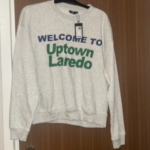 Grå sweatshirt Uptown Laredo print - Grå sweatshirt med trycket 'WELCOME TO Uptown Laredo' i blått och grönt framtill. Klassisk rund halsringning och ribbade muddar vid ärmslut och nederkant. Perfekt för en chill och avslappnad stil.