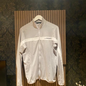 Ljusgrå Tröja från J.Lindeberg ”Jarvis” - Snygg ljusgrå ”Golf” tröja från J.Lindeberg med vit horisontell detalj över bröstet och ryggen. Tröjan har hel dragkedja framtill, hög krage och  logga på bröstet. Perfekt för dig som gillar stilren och sportig look. Fungerar som gym tröja eller vardaglig tröja. 