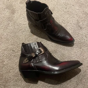 Svart/bruna äkta cowboy boots med spänne - Snygga svarta/bruna äkta cowboy boots i skinn med spetsig tå och dekorativa sömmar. Skorna har ett coolt metallspänne på sidan och låg klack. Perfekta för dig som vill sticka ut med western-vibe och detaljerad finish. Made in Portugal. Köpt sacond hand för ca 3000 kr då det är Vintage. Supercoola❤️