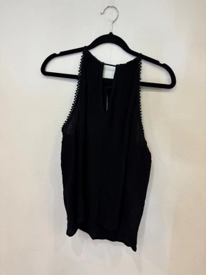 Svart ärmlös linnetopp med kantdetalj - Svart ärmlös linnetopp med rund halsringning och dekorativa små bollar längs ärmhålen. Toppen har en lätt och luftig känsla, knäppning i nacken och en liten öppning framtill. Perfekt för en stilren och enkel look.