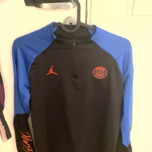 Jordan PSG svart/blå half zip tröja - Svart och blå långärmad tröja från Jordan PSG med röd logga och Paris Saint-Germain emblem på bröstet. Slim fit modell med half zip framtill och coolt tryck på ärmen. Perfekt för dig som gillar sportig stil och fotboll.