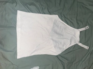 Vit ärmlös linnetopp med knappar - Säljer en stilren vit linnetopp med raka linjer och breda axelband som knäpps med metallknappar. Toppen är ärmlös och har en enkel, clean look som passar perfekt till sommaren. Materialet är lätt och har en diskret struktur.