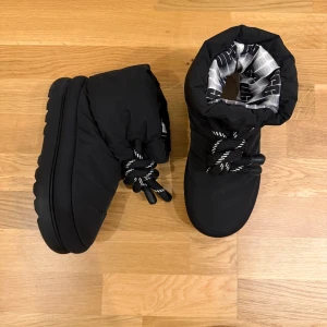 Svarta skor från UGG, strl 39 - Skor från UGG i svart med tjock, räfflad platt sula och rund tå. Yttermaterialet är syntet och insidan har ett mönstrat foder i grått och svart och innersulan i päls. Skorna har snörning med grova svarta och vita snören och dragsko upptill. Perfekta för kalla vinterdagar.