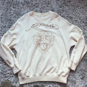 Beige Ed Hardy sweatshirt tigerprint Storlek M - Snygg beige sweatshirt från Ed Hardy i storlek M, tillverkad i 100% bomull. Framsidan har ett stort tigertryck och Ed Hardy-logga i beige, medan baksidan har loggan tryckt upptill. Tröjan har rund hals och långa ärmar med ribbade muddar och två dolda fickor. Knappt använd! Rekommenderar starkt