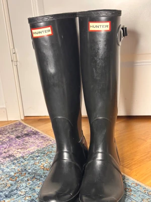 Svarta Hunter stövlar i gummi - Klassiska svarta höga Hunter stövlar i gummi med justerbart spänne på sidan och logga framtill. Perfekta för regniga dagar och festivaler. Snygg, tidlös design som funkar till många olika outfits.