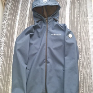 Blå vindjacka från Sail Racing - Storlek 176 men passar som 170 och xs. Snygg blå vindjacka från Sail Racing med huva och dragkedja. Jackan har en stilren design med logga på bröstet och är fodrad med mjuk fleece på insidan. Perfekt för dig som gillar sportig och clean look. Justerbar huva och praktiska fickor.             