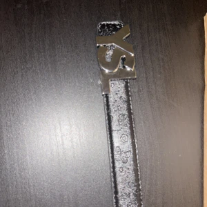 Svart YSL bälte med silverdetaljer - Helt ny Snyggt svart bälte från YSL med en stor, blank silverspänne i form av YSL-loggan. Bältet är i skinn och har präglade fjärilar och blommor längs hela remmen för en unik touch. Perfekt för att lyfta vilken outfit som helst. Det är för både herr och damer. Pris kan diskuteras !