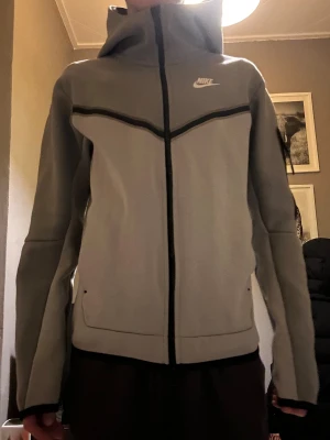 Grå Niketech tröja med luva - Snygg grå zip hoodie från Nike med svart dragkedja och coolt svart detaljband över bröstet. Tröjan har luva, två sidofickor och en stilren look. Perfekt för dig som gillar sportig stil.